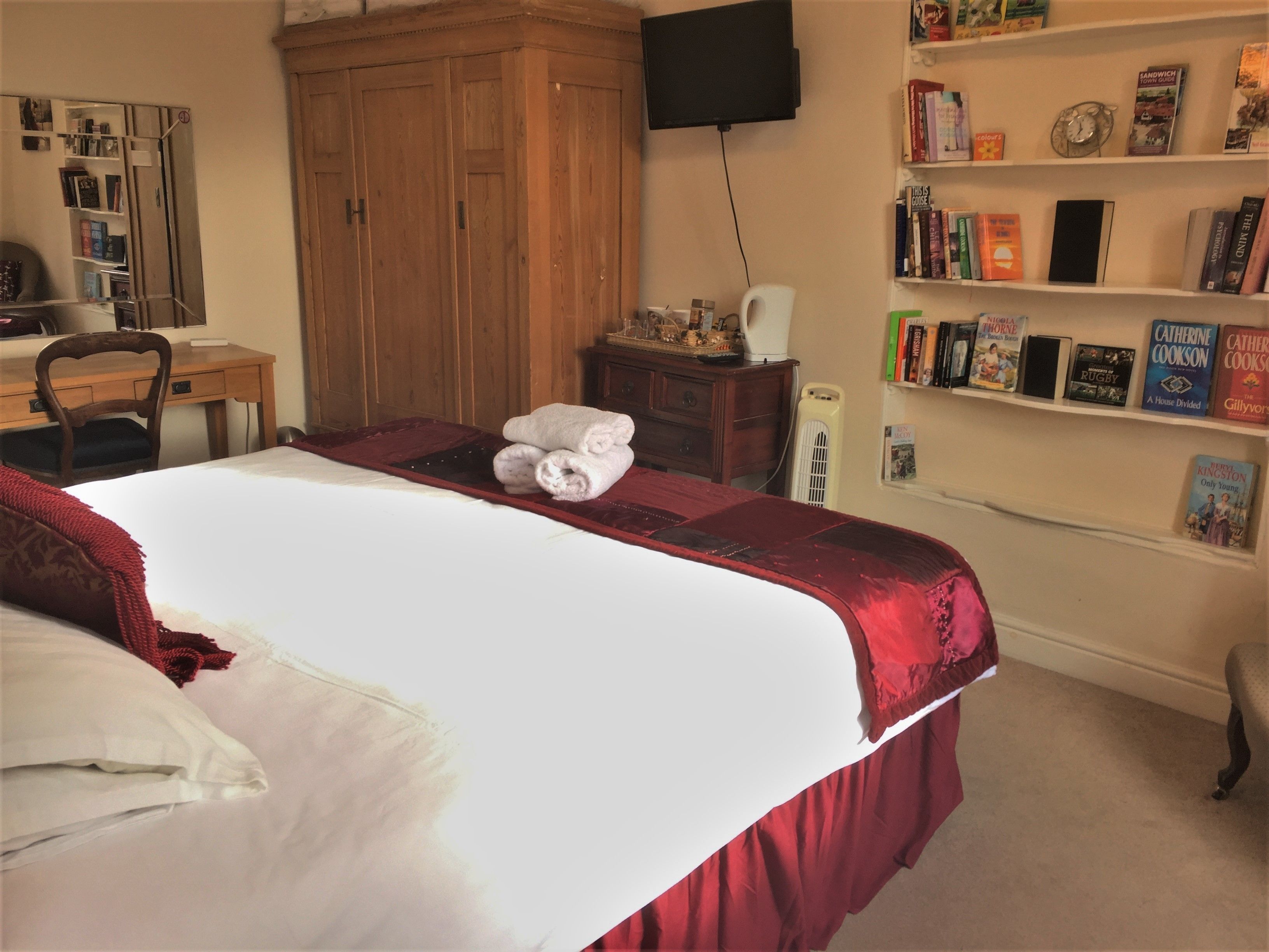 A neatly made bed with a red and white bedspread, a wooden wardrobe, a desk with a chair, a bookshelf with various books, and a television mounted on the wall.