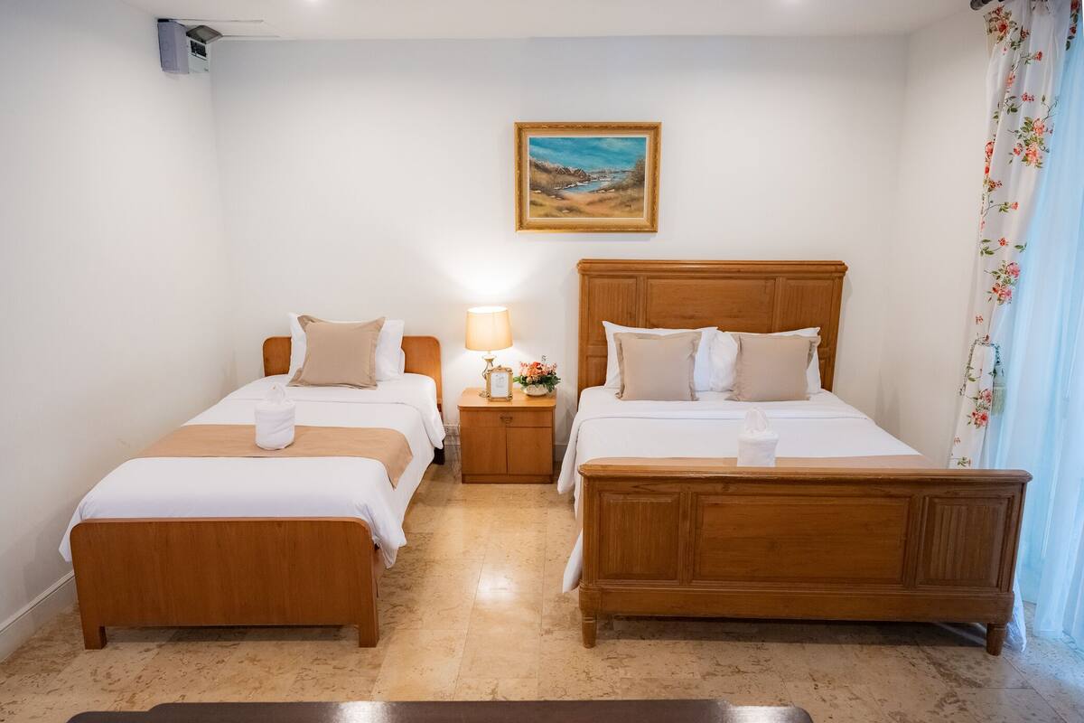 A hotel room with two single beds, a wooden headboard, a nightstand with a lamp, a painting on the wall, and a floral curtain.