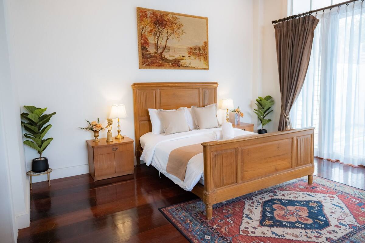 A bedroom with a wooden bed, bedside tables, a nightstand, a large painting, and a window with curtains.