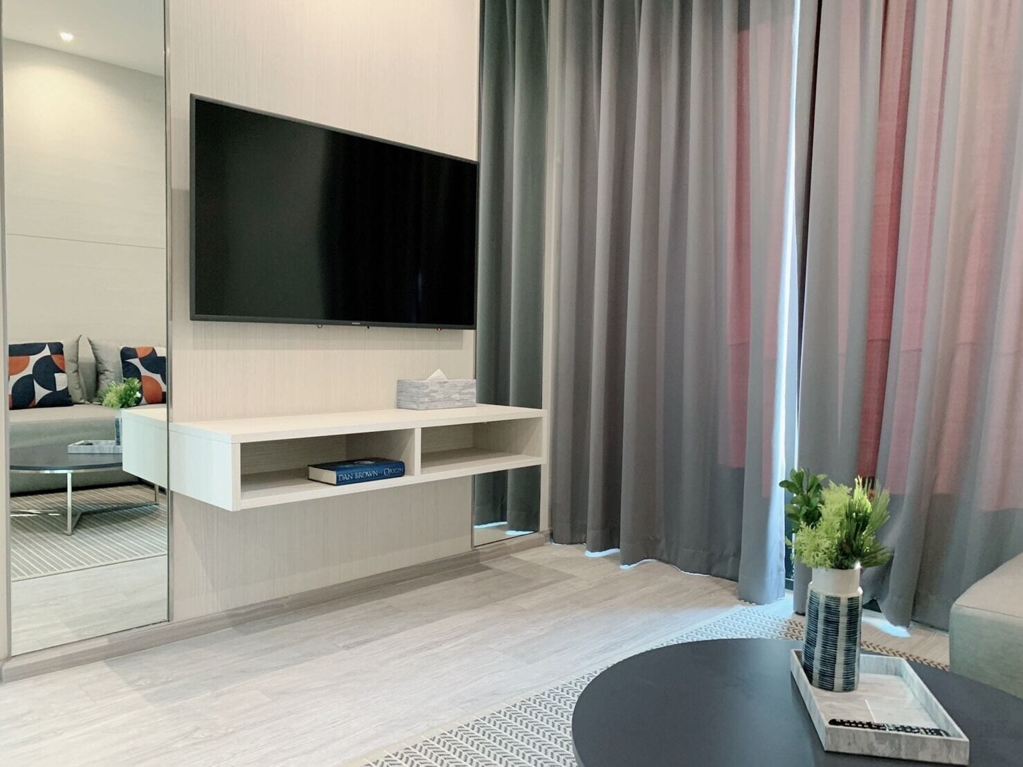A modern living room with a flat-screen TV mounted on a wall, a white entertainment unit, a glass coffee table, and a grey sofa with patterned cushions.