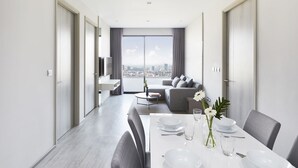 Un comedor moderno con una mesa blanca dispuesta para cuatro personas, un sofá al fondo y vistas a la ciudad a través de amplios ventanales.