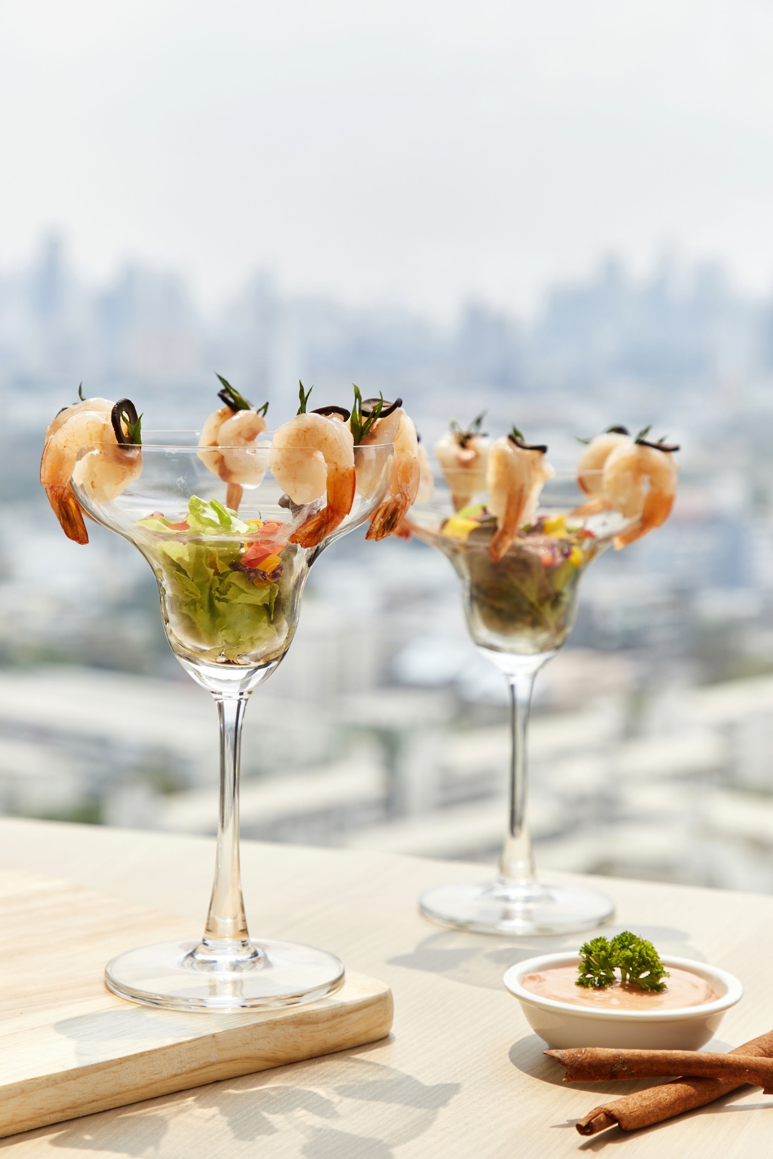 Dos vasos de martini con brochetas de camarón y una ensalada, sobre una mesa de madera con vista a la ciudad.