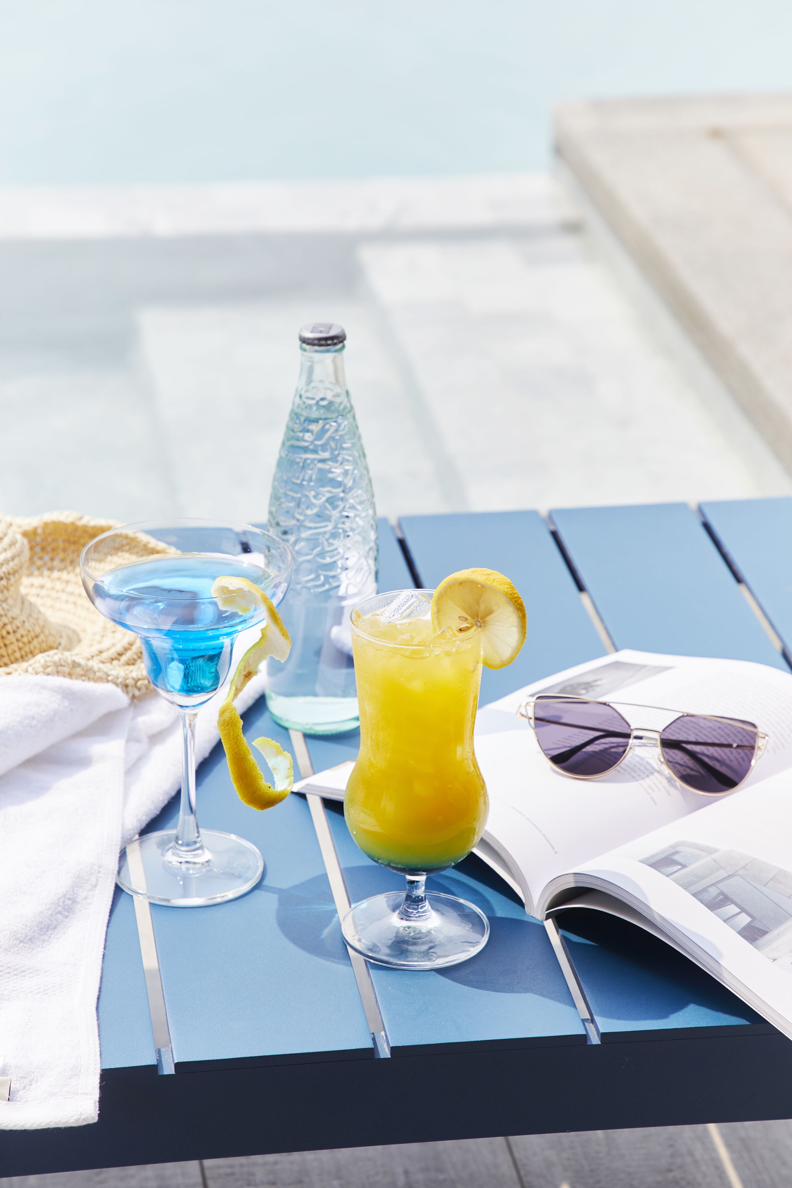 Une table avec deux boissons, une bouteille et des lunettes de soleil, posée sur une nappe bleue près d’une piscine.