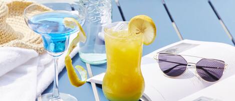 Une table avec deux boissons, une bouteille et des lunettes de soleil, posée sur une nappe bleue près d’une piscine.