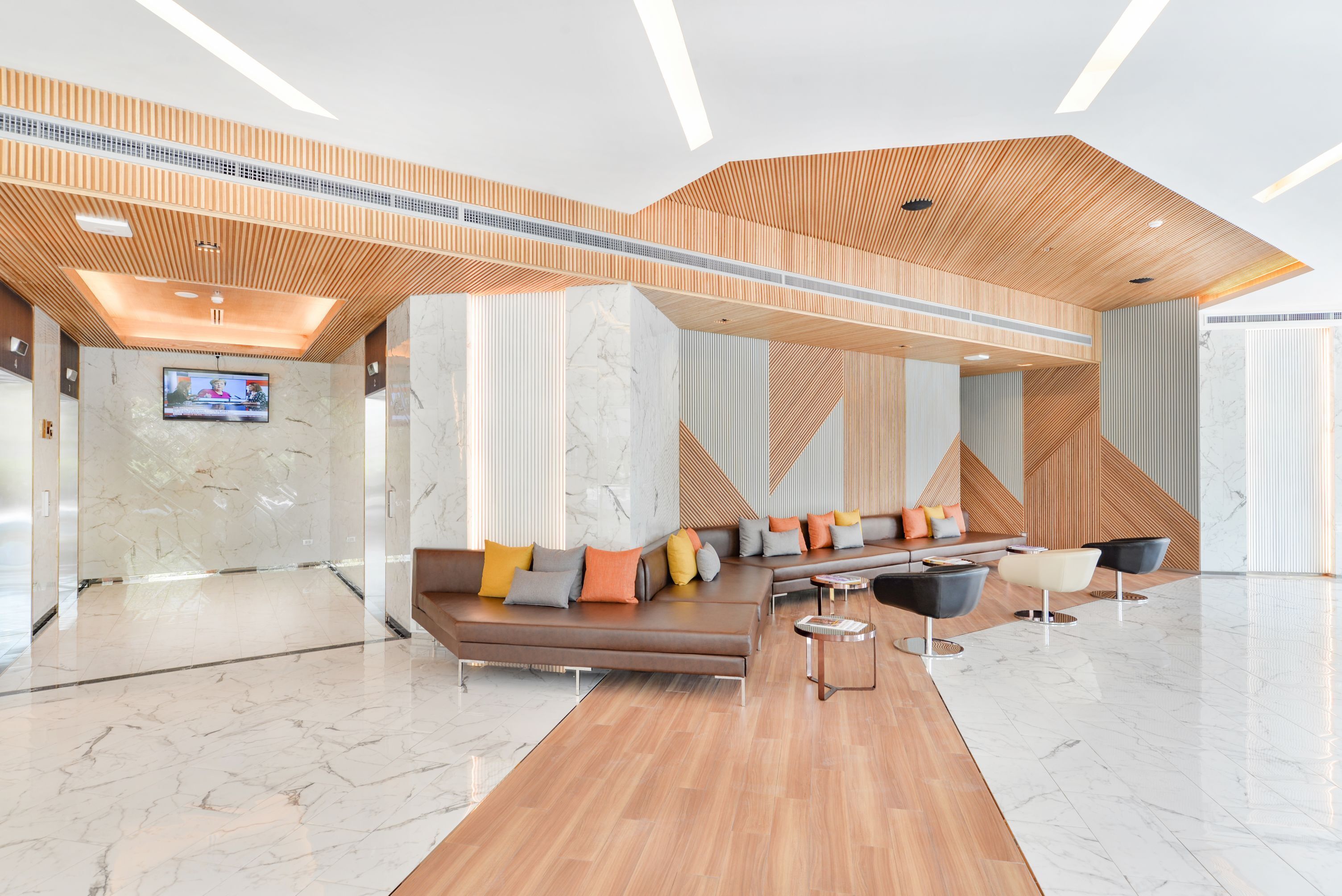 A modern lobby with a wooden ceiling, a marble reception desk, a seating area with sofas and chairs, and a flat-screen TV on the wall.