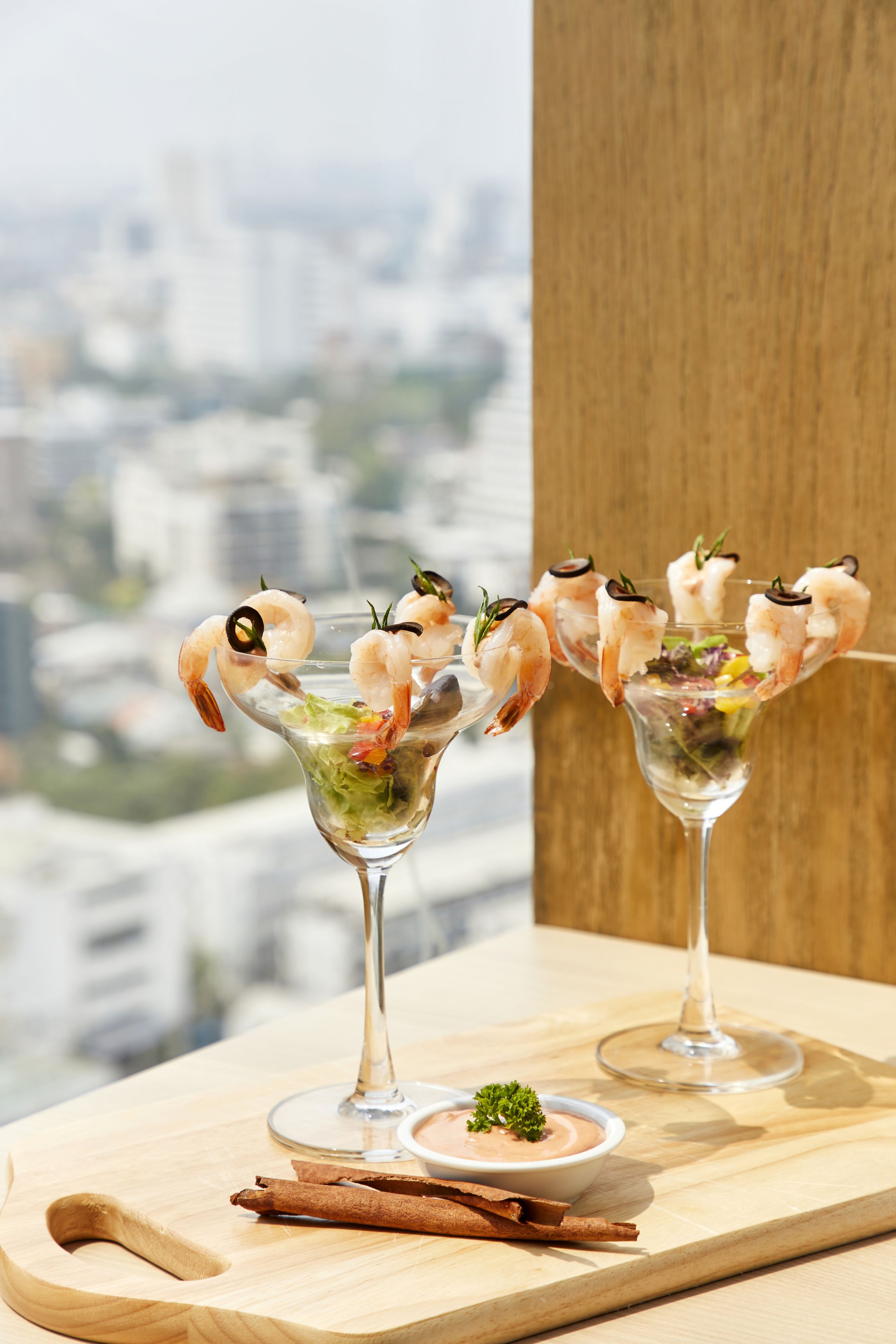 Two martini glasses with shrimp cocktails on a wooden board, with a cityscape view in the background.