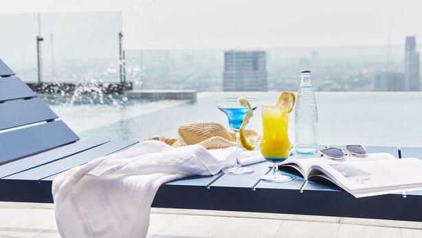 Escena junto a la piscina con sillones de descanso, una toalla, un cóctel, una botella y gafas de sol sobre una mesa.