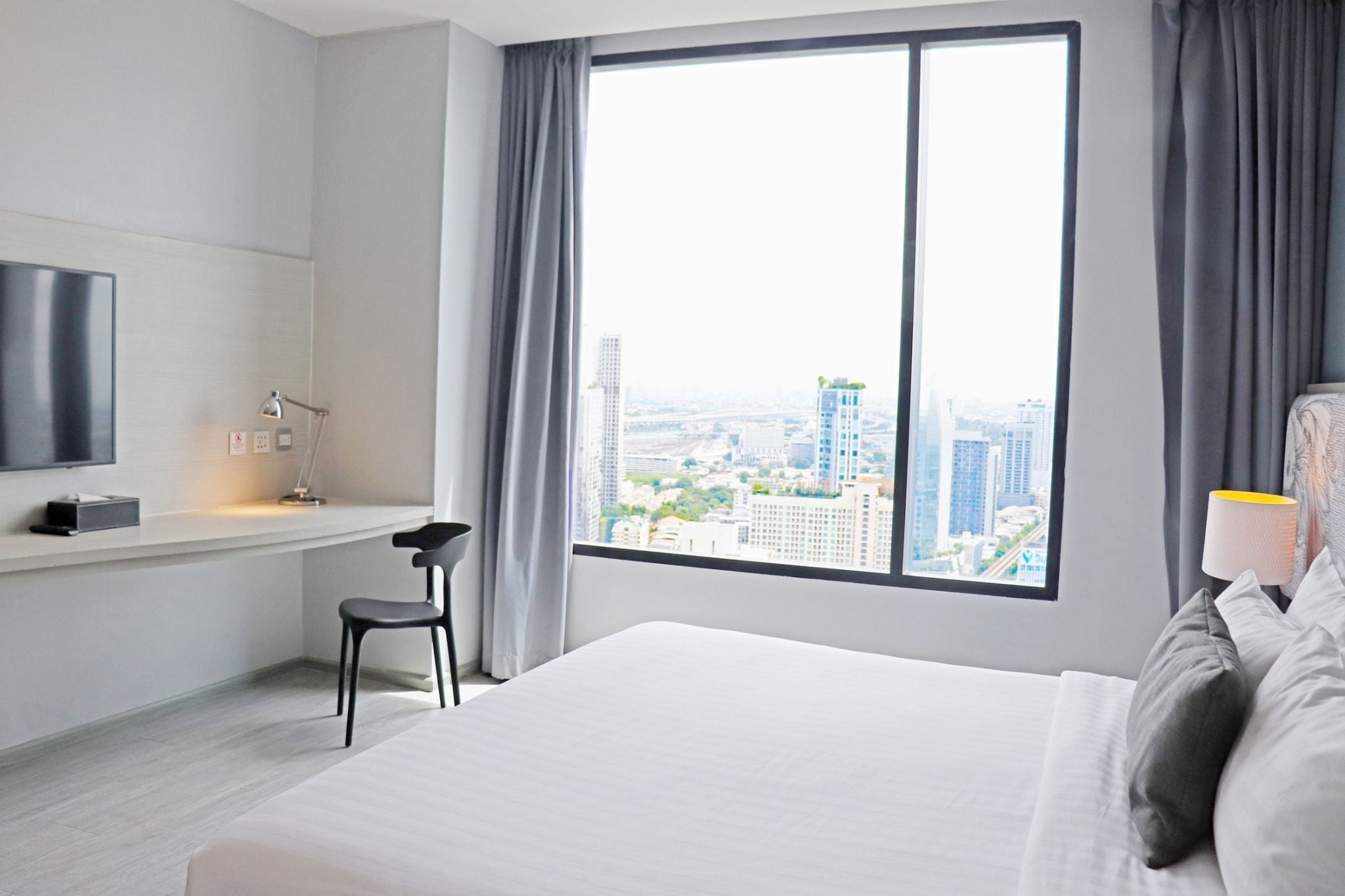 A modern hotel room with a large window offering a city view, a bed, a desk with a chair, and a TV mounted on the wall.