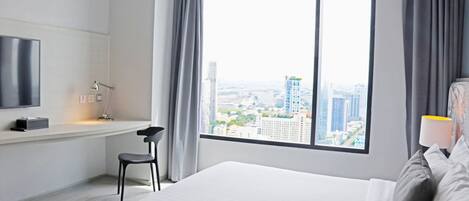 A modern hotel room with a large window offering a city view, a bed, a desk with a chair, and a TV mounted on the wall.