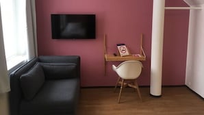 A room with a dark sofa, a wooden desk with a chair, a television on the wall, and a window with curtains. - Place to Sleep Hotel Rauma (Rauma)