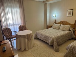 Habitación de hotel con cama, una mesa redonda, dos sillas, una mesita de noche y una lámpara fijada a la pared.