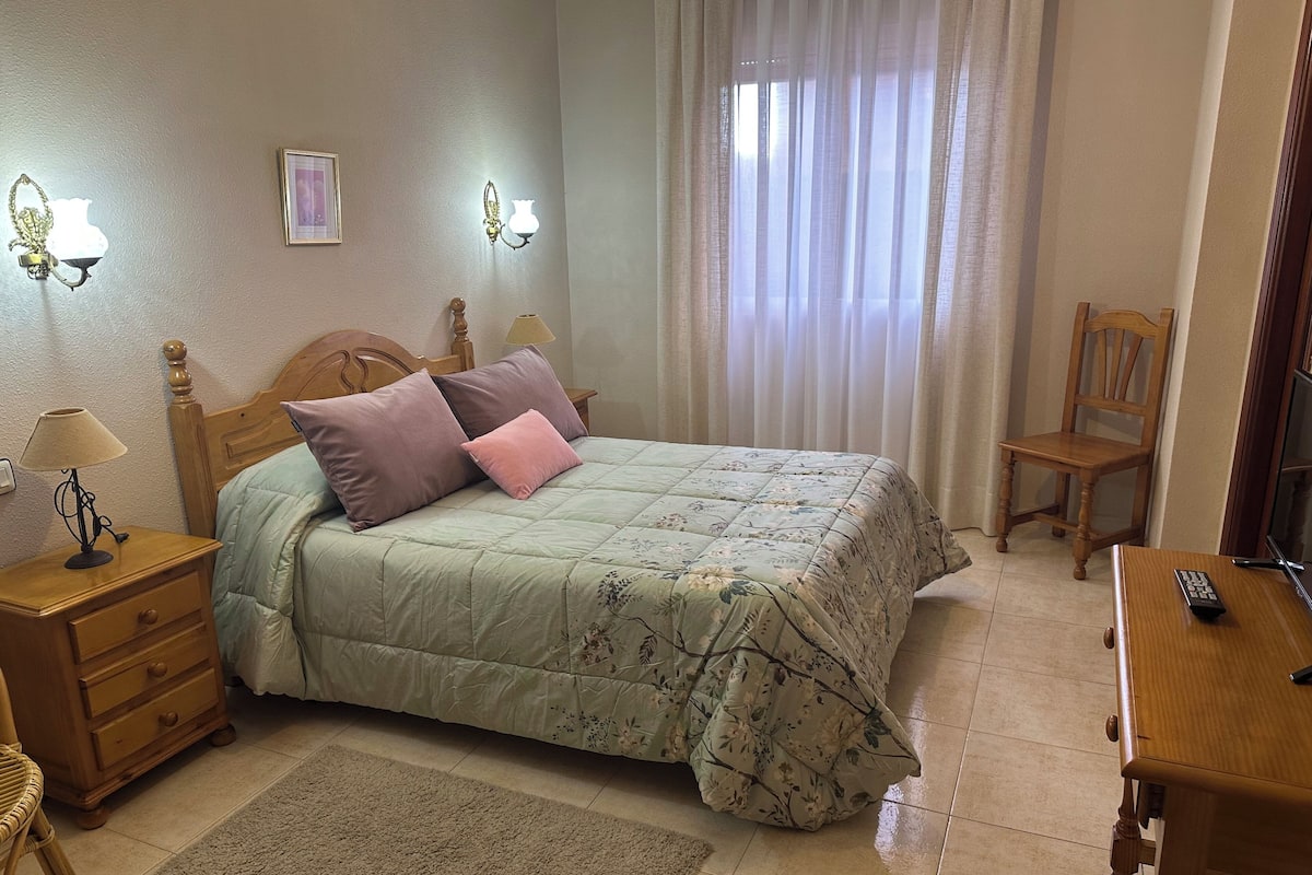 Ein Schlafzimmer mit einem Bett, zwei Nachttischlampen, einem Holznachttisch, einem Sessel und einem Schreibtisch mit Fernbedienung.