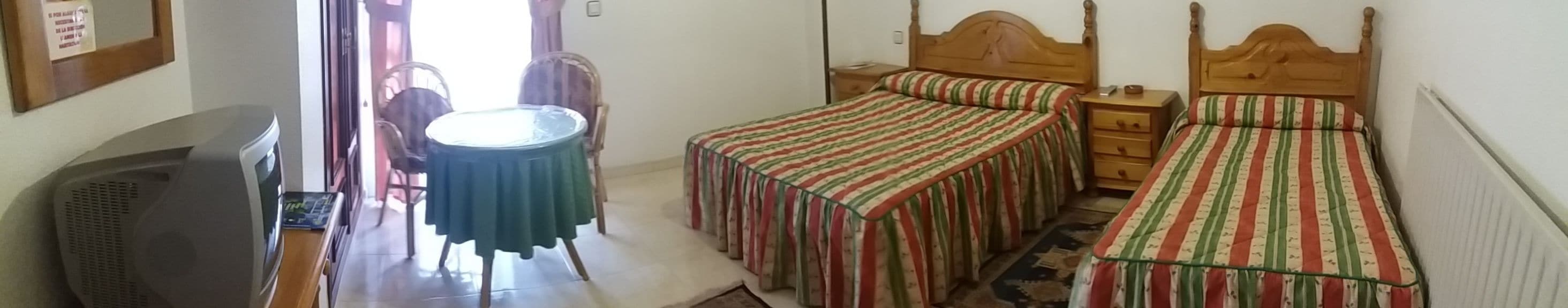 Una habitación con una cama, una mesita de noche, una silla, una pequeña mesa con mantel verde y un televisor.