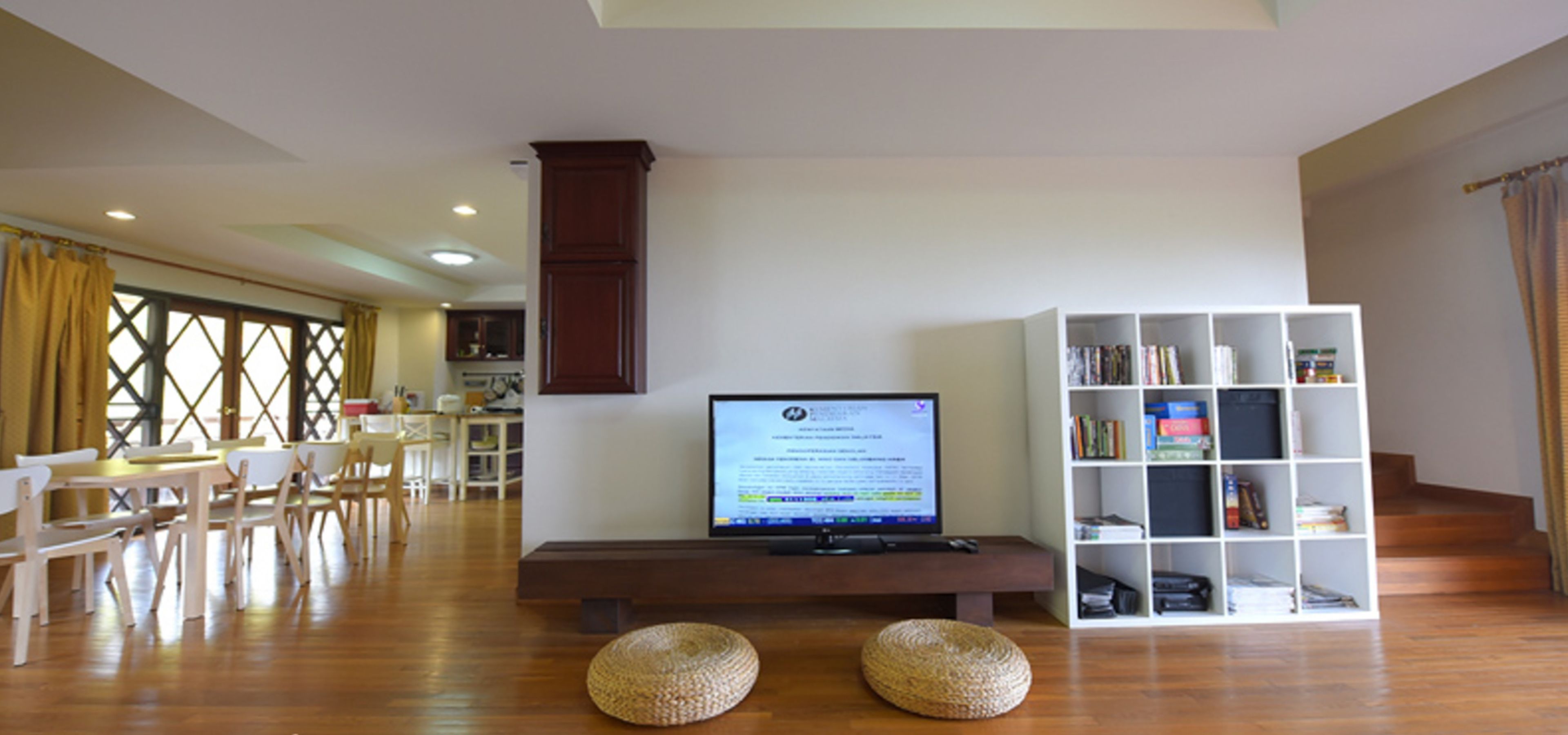 A modern living room with a flat-screen TV on a wooden bench, a bookshelf, and a dining area with chairs and a table.