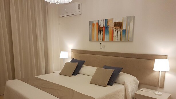 Une chambre d’hôtel avec un grand lit, deux tables de chevet avec des lampes, un climatiseur fixé au mur et une peinture abstraite aux couleurs vives au-dessus du lit.
