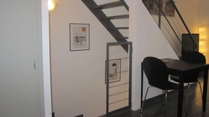 Una escalera con barandilla negra que lleva a un rellano. Hay un cuadro en la pared y una pequeña mesa con sillas.