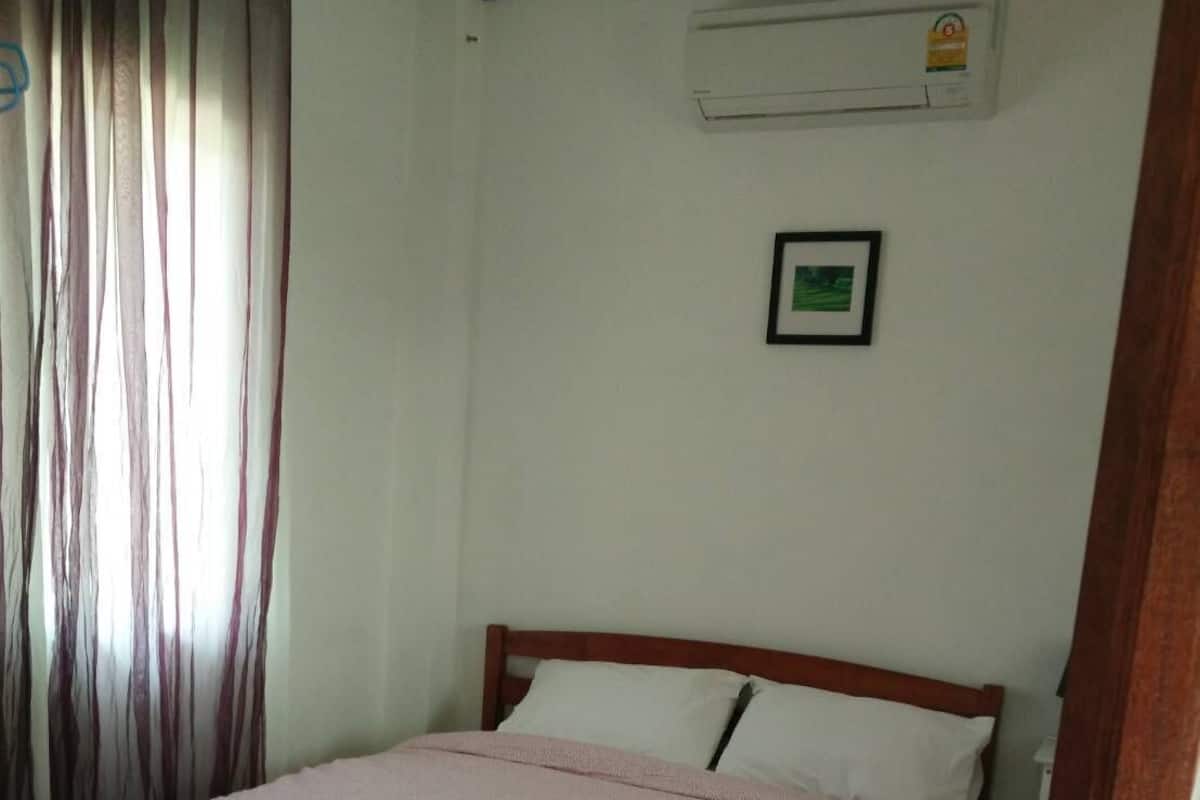 A single bed with a wooden frame and a pink blanket, a white wall with a framed picture, and a window with curtains.