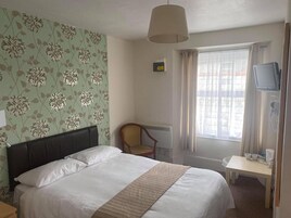 Una habitación de hotel con cama, una silla, escritorio con televisor y ventana con cortinas.