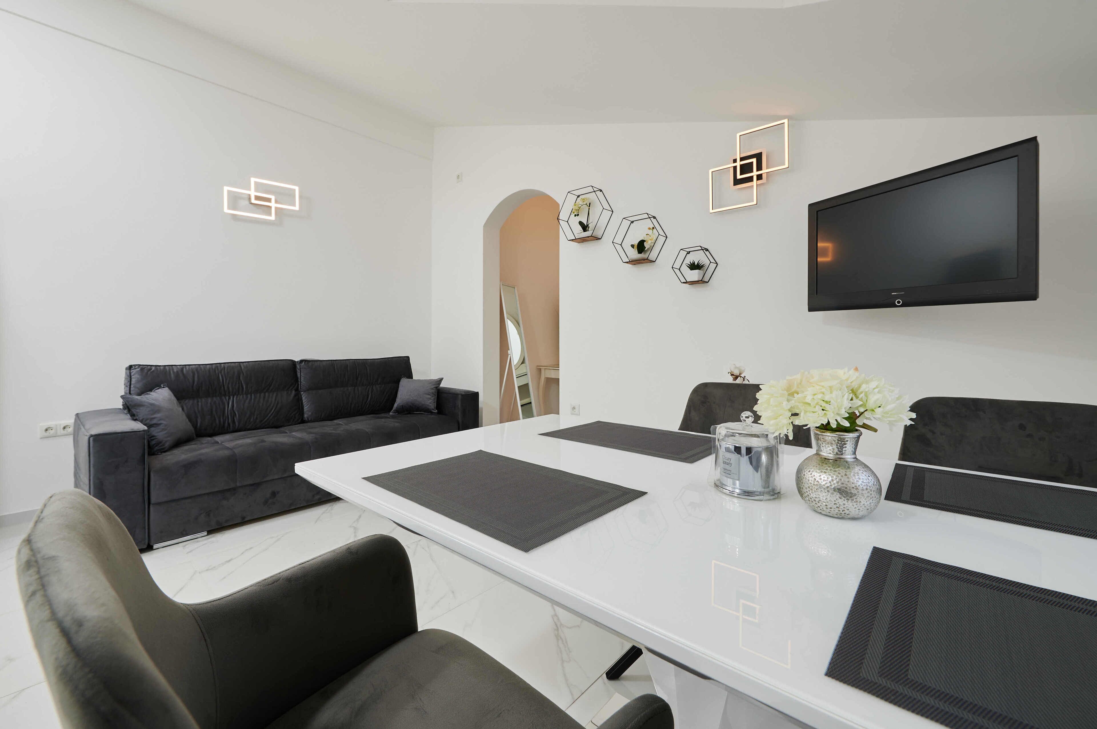 A modern living room with a white table, black sofas, and a mounted TV.