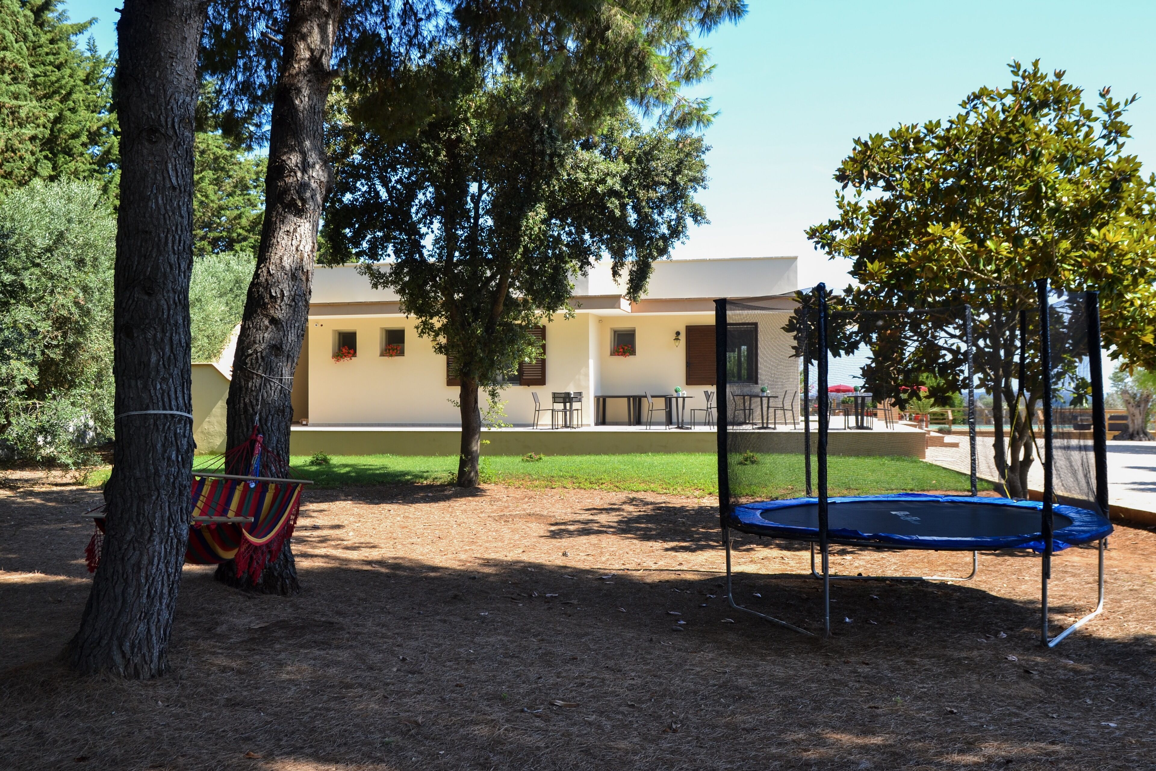 A playground with a trampoline and a swing set, surrounded by trees and a building with multiple windows.