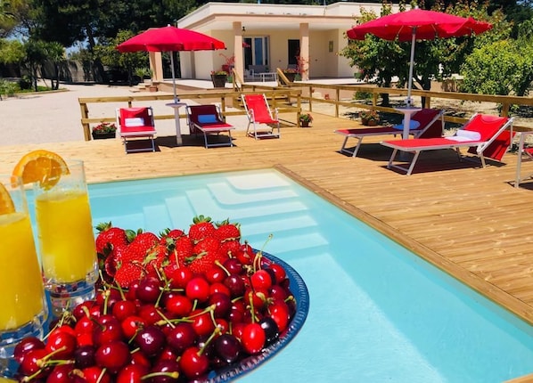 Un área junto a la piscina con sillones de descanso, un cuenco de frutas con cerezas y fresas, y sombrillas rojas.