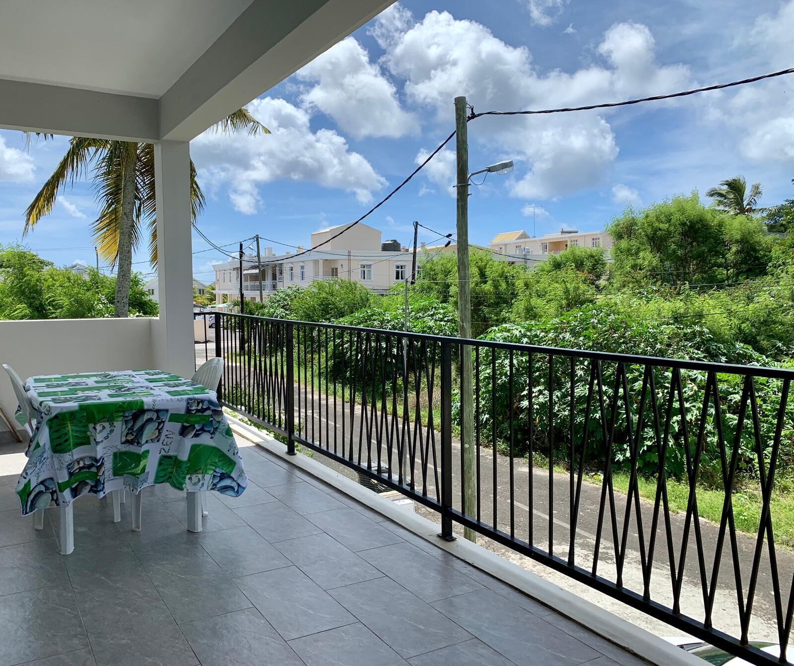 A balcony with a table covered in a green and white tablecloth, a black metal railing, and a view of a street with buildings and greenery.