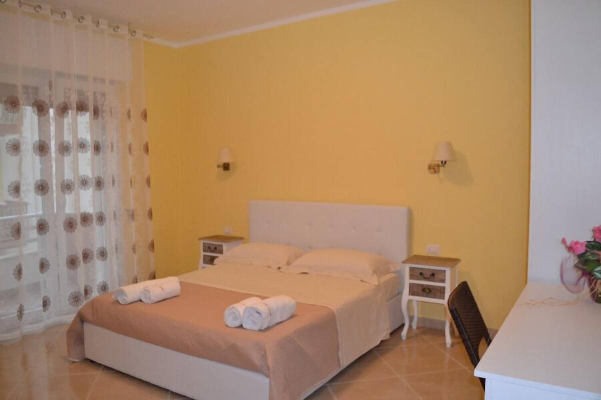 A neatly made bed with a brown bedspread and two rolled towels, a bedside table with a lamp, and a small chair.