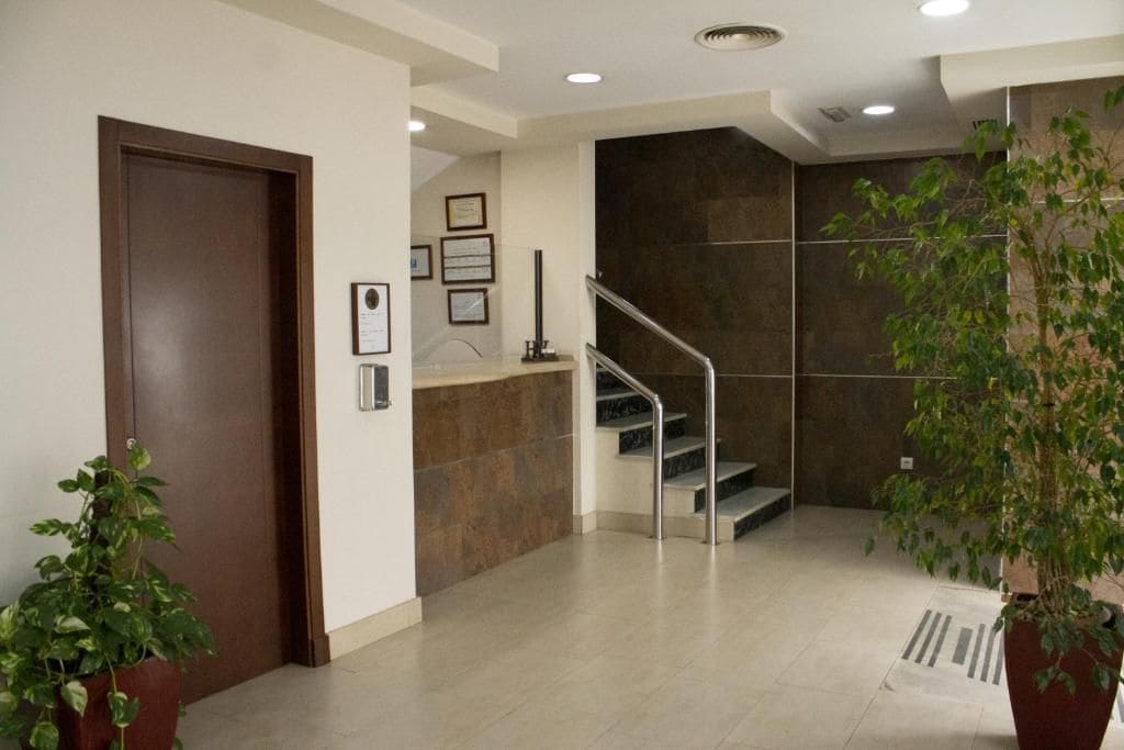 Un vestíbulo moderno con puerta de madera, mostrador de recepción, escalera y plantas en macetas.