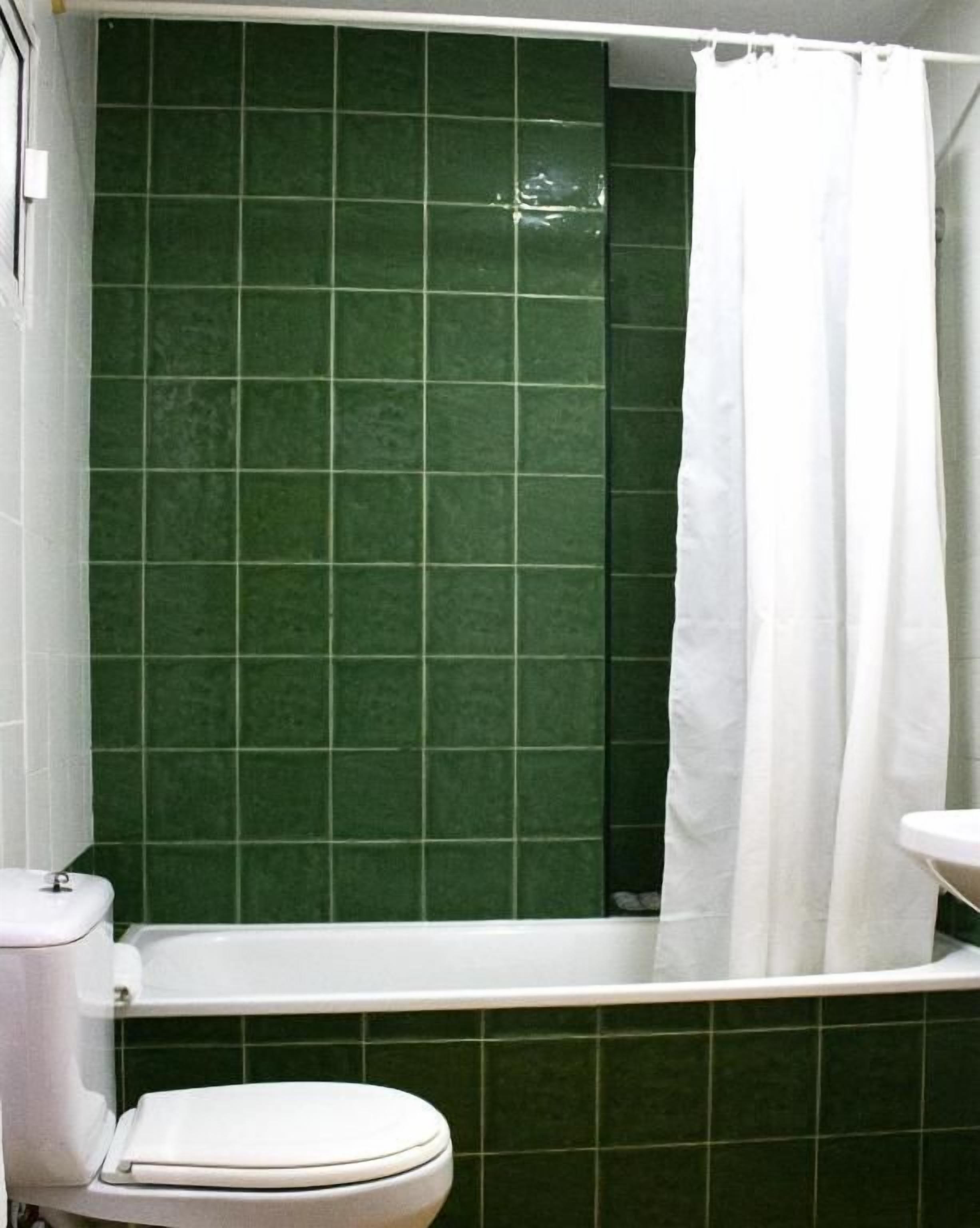 Un baño con pared de azulejos verdes, un inodoro blanco y una bañera blanca con cortina de ducha blanca.