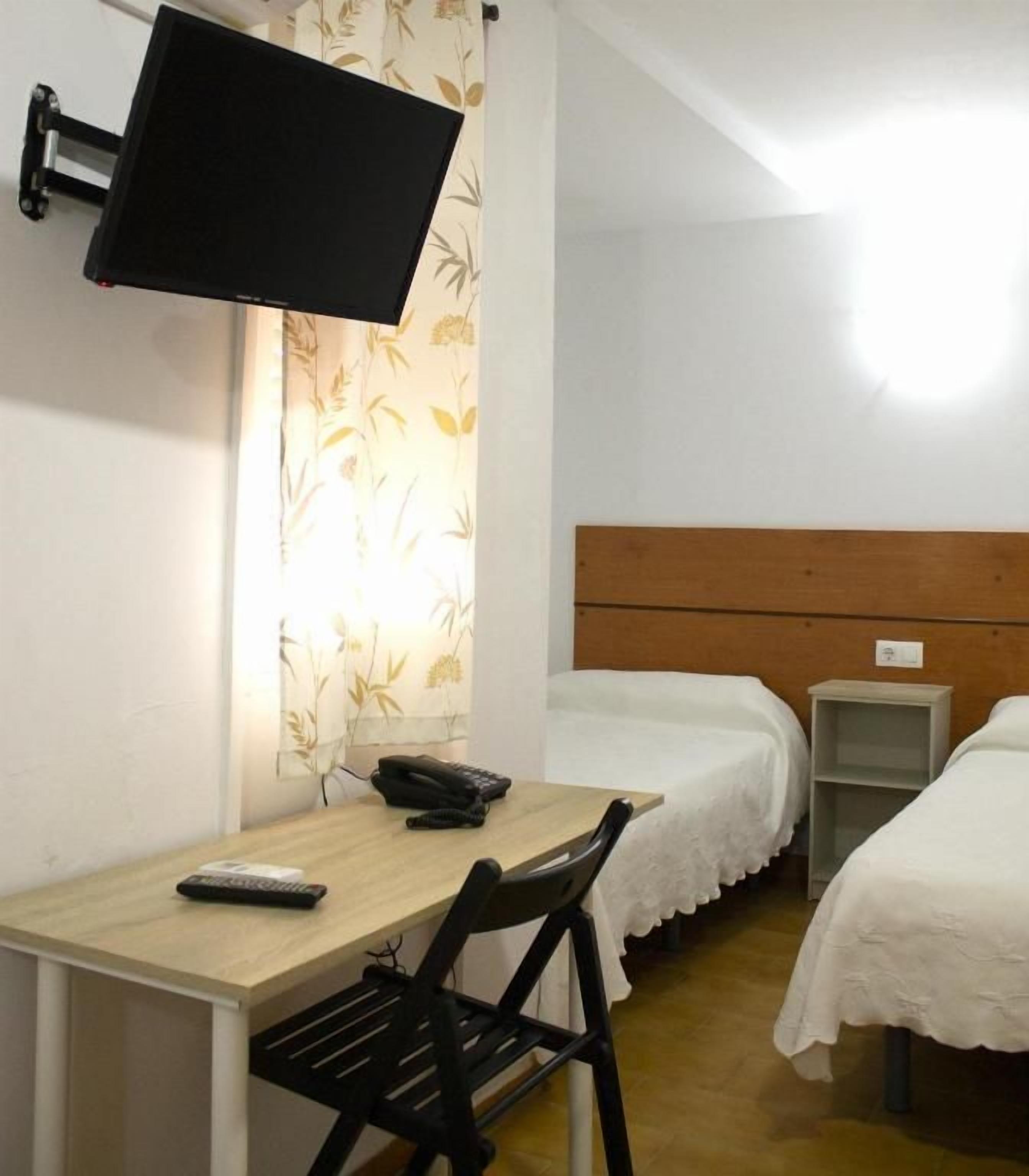 Una habitación con cama, escritorio, silla, televisor montado en la pared y una pequeña estantería.