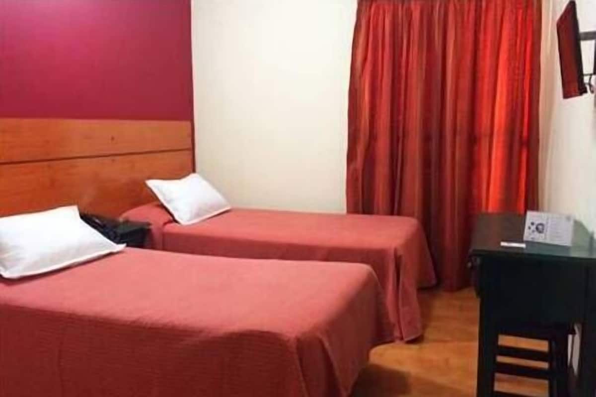 Une chambre d’hôtel avec deux lits simples, une tête de lit en bois, un bureau avec une chaise et un rideau rouge.
