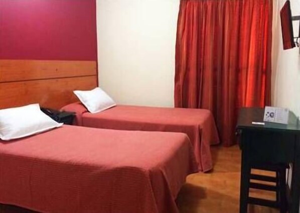 A hotel room with two single beds, a wooden headboard, a desk with a chair, and a red curtain.
