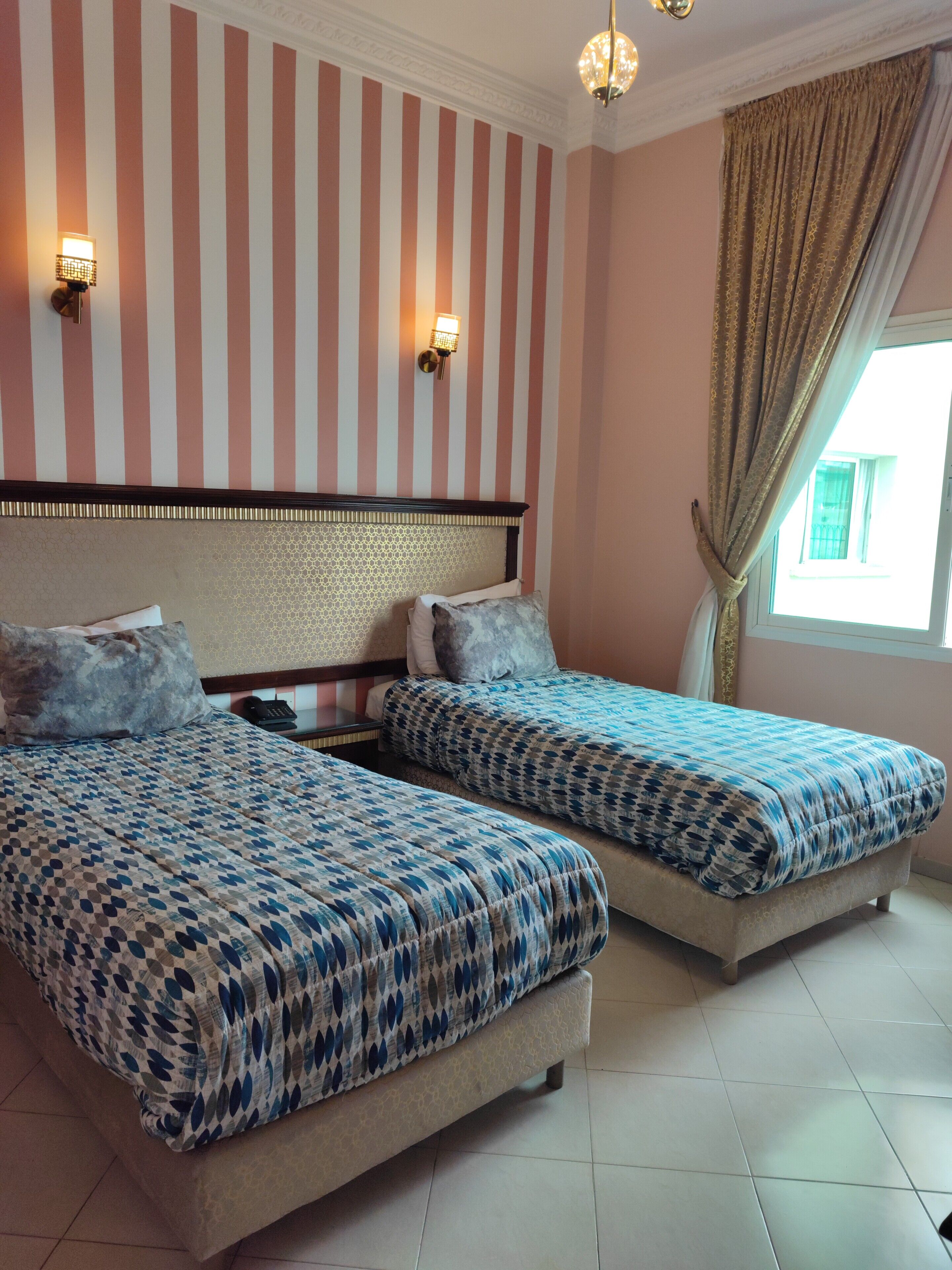 A hotel room with two beds, striped wallpaper, a patterned headboard, a telephone, and a window with curtains.