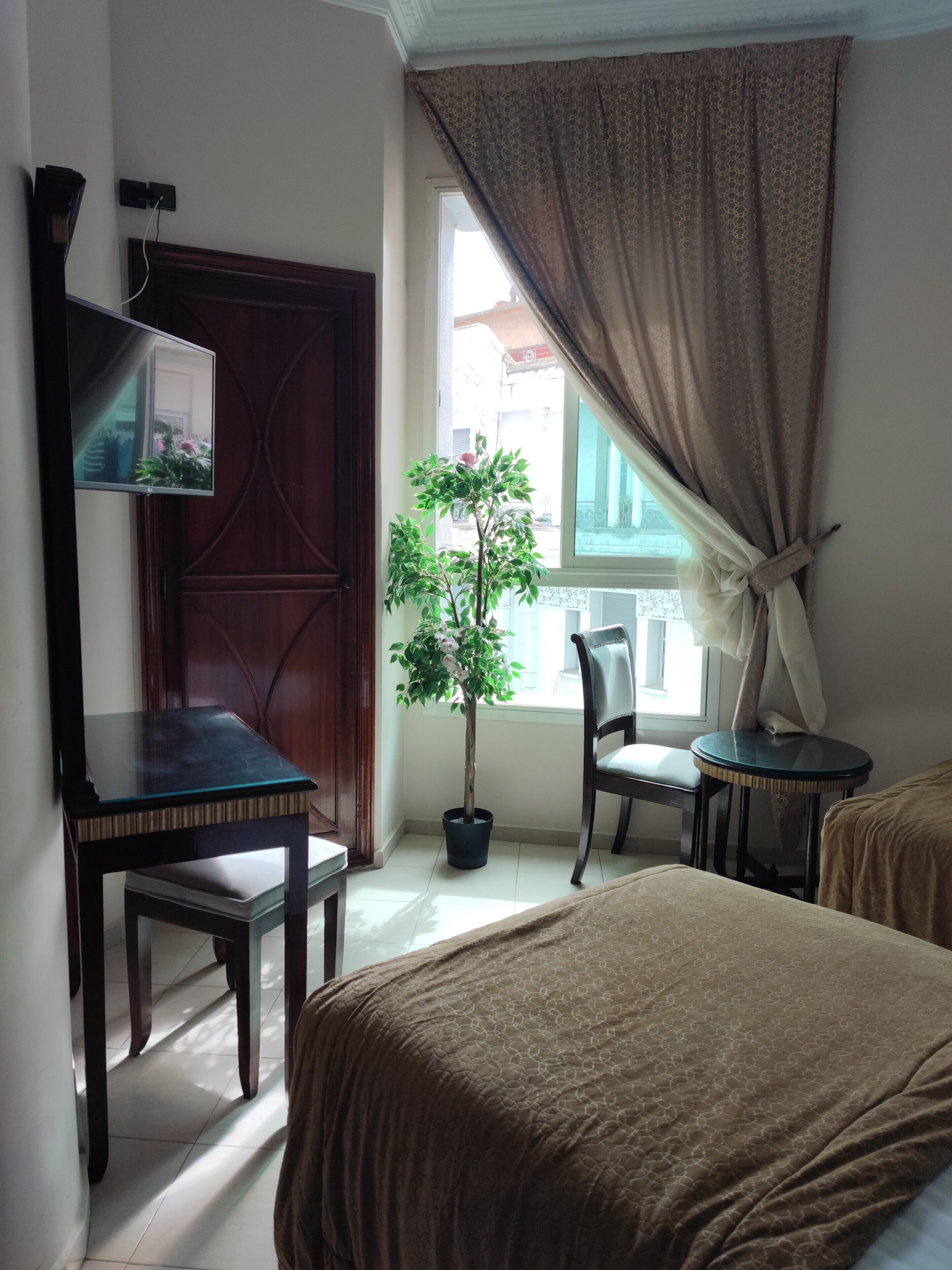 A room with a bed, a desk, a chair, a small table, a potted plant, and a window with curtains.