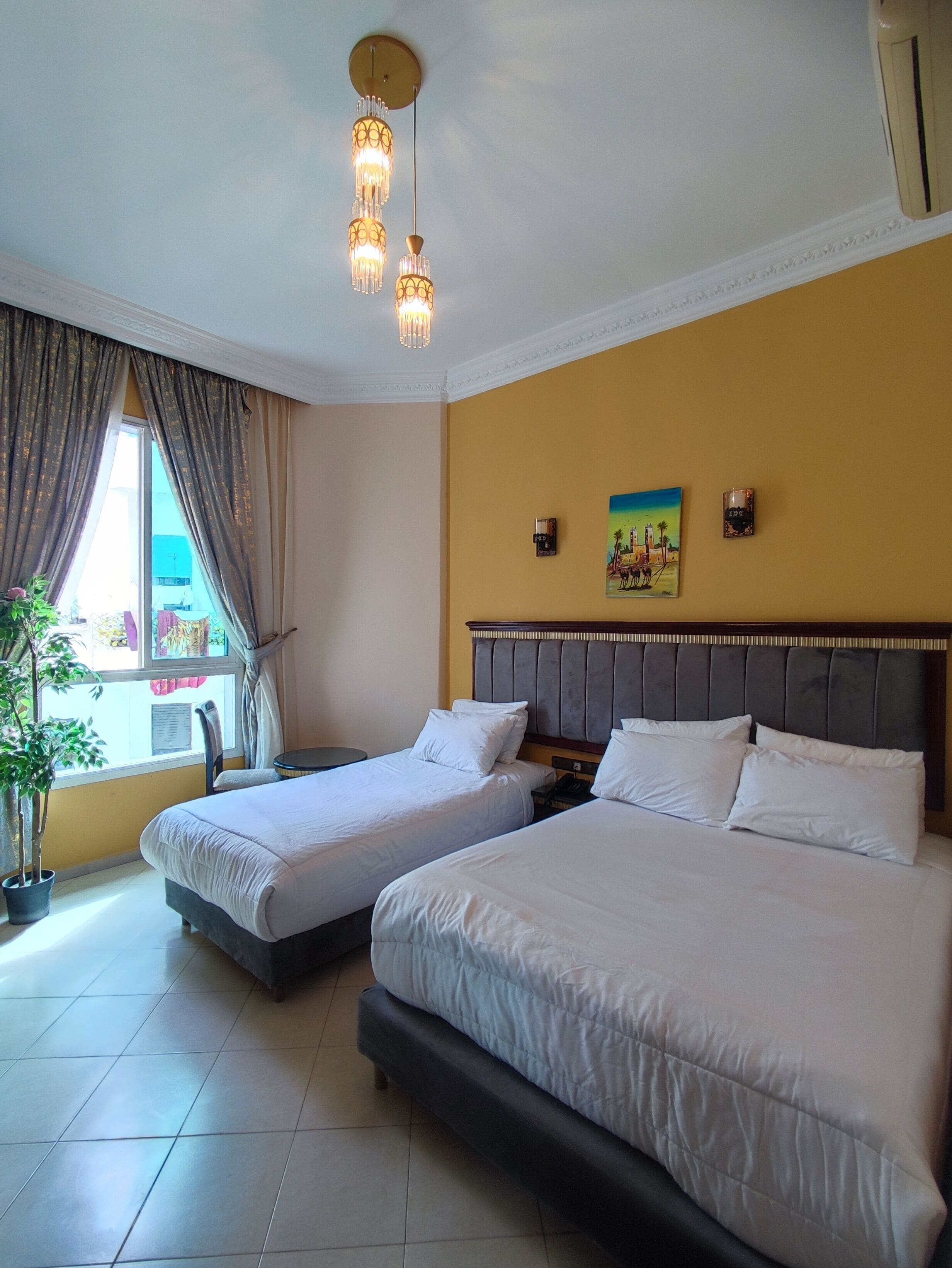 A hotel room with two beds, a large window with curtains, a painting on the wall, and a ceiling light fixture.