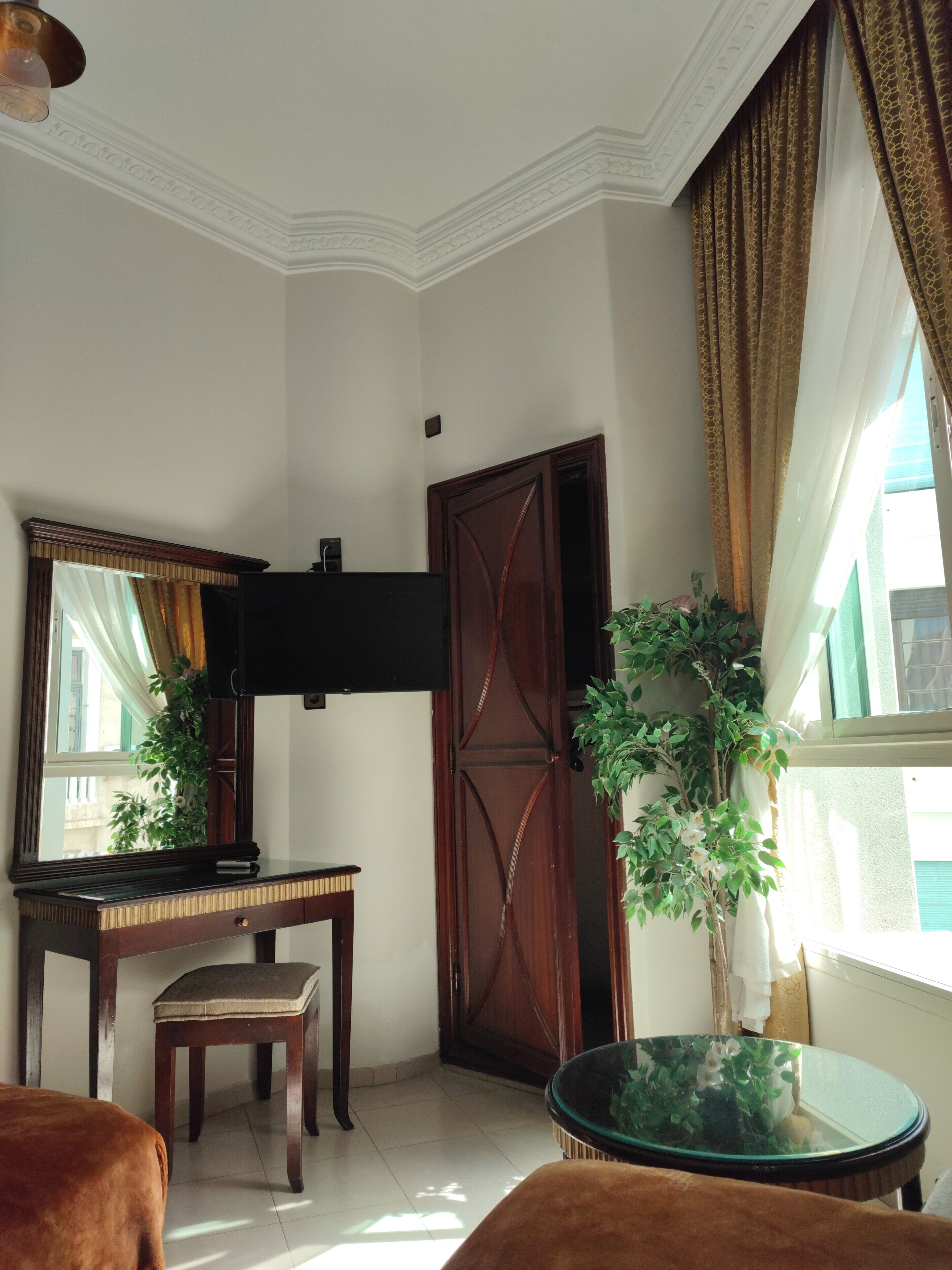 Ein Zimmer mit Schreibtisch aus Holz, einem Stuhl, einem an der Wand befestigten Fernseher, einem großen Spiegel, einer Holztür und einem Blumentopf am Fenster.