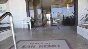 L’entrée d’un hôtel, d’où l’on aperçoit clairement l’intérieur, avec une chaise blanche et une enseigne indiquant « EDIFICIO SAN REMO ».