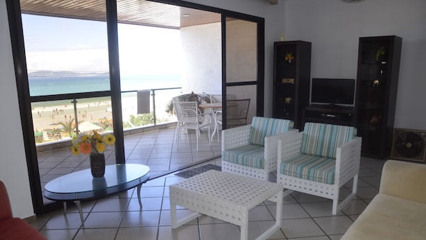 Un salon avec un balcon donnant sur une plage, des meubles blancs et un meuble pour la télévision.