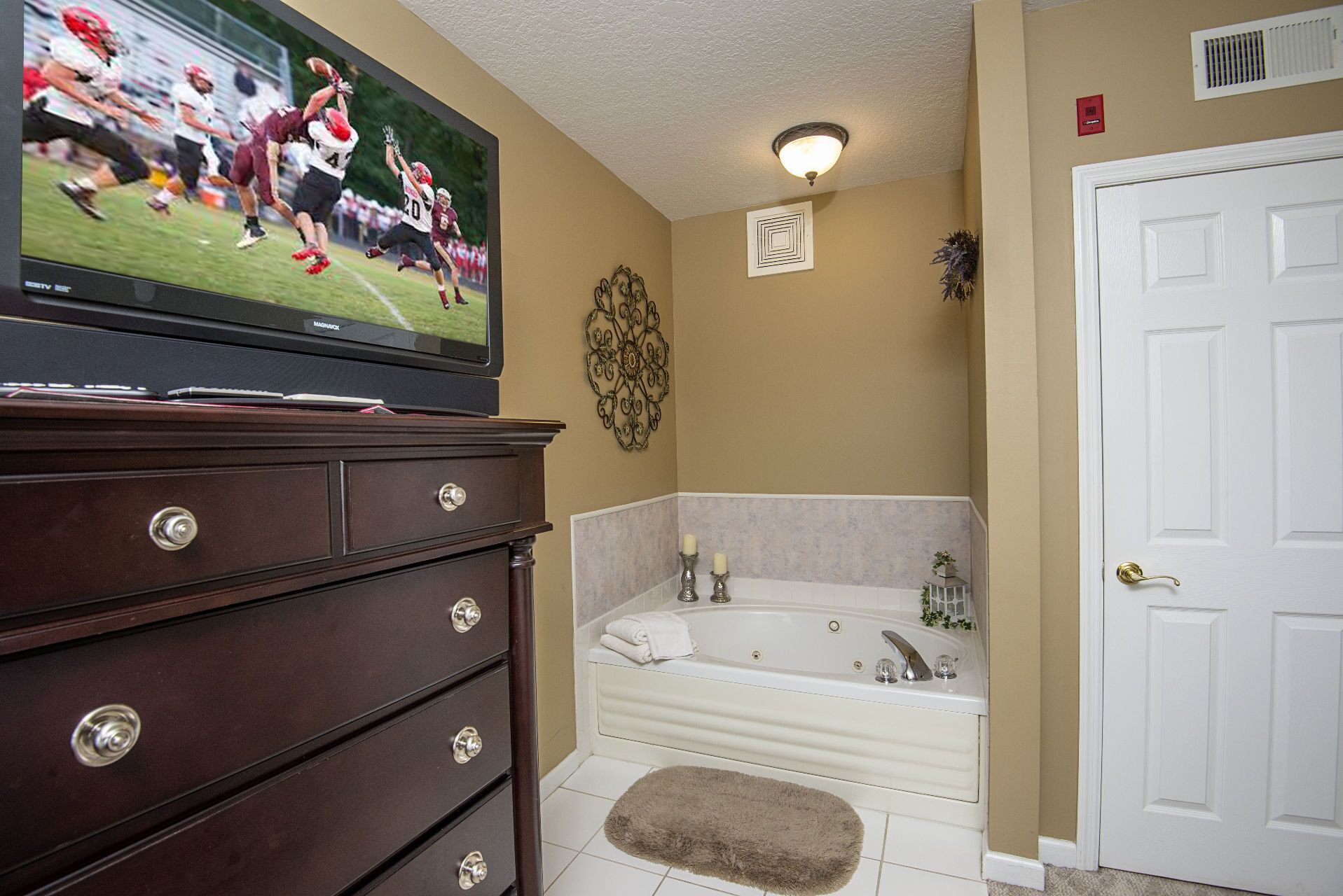 A bathroom with a bathtub, a vanity with drawers, a television displaying a sports game, and a door.