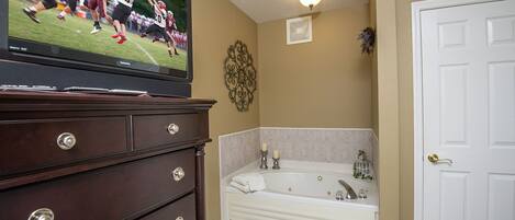 A bathroom with a bathtub, a vanity with drawers, a television displaying a sports game, and a door.