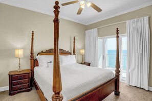 A four-poster bed with a wooden frame, two bedside tables with lamps, and a view of the ocean through a balcony with white curtains.