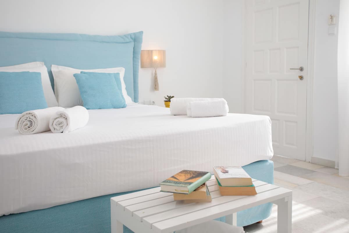 Una cama bien hecha con ropa de cama azul y blanca, una mesita de noche con libros, y una puerta al fondo.