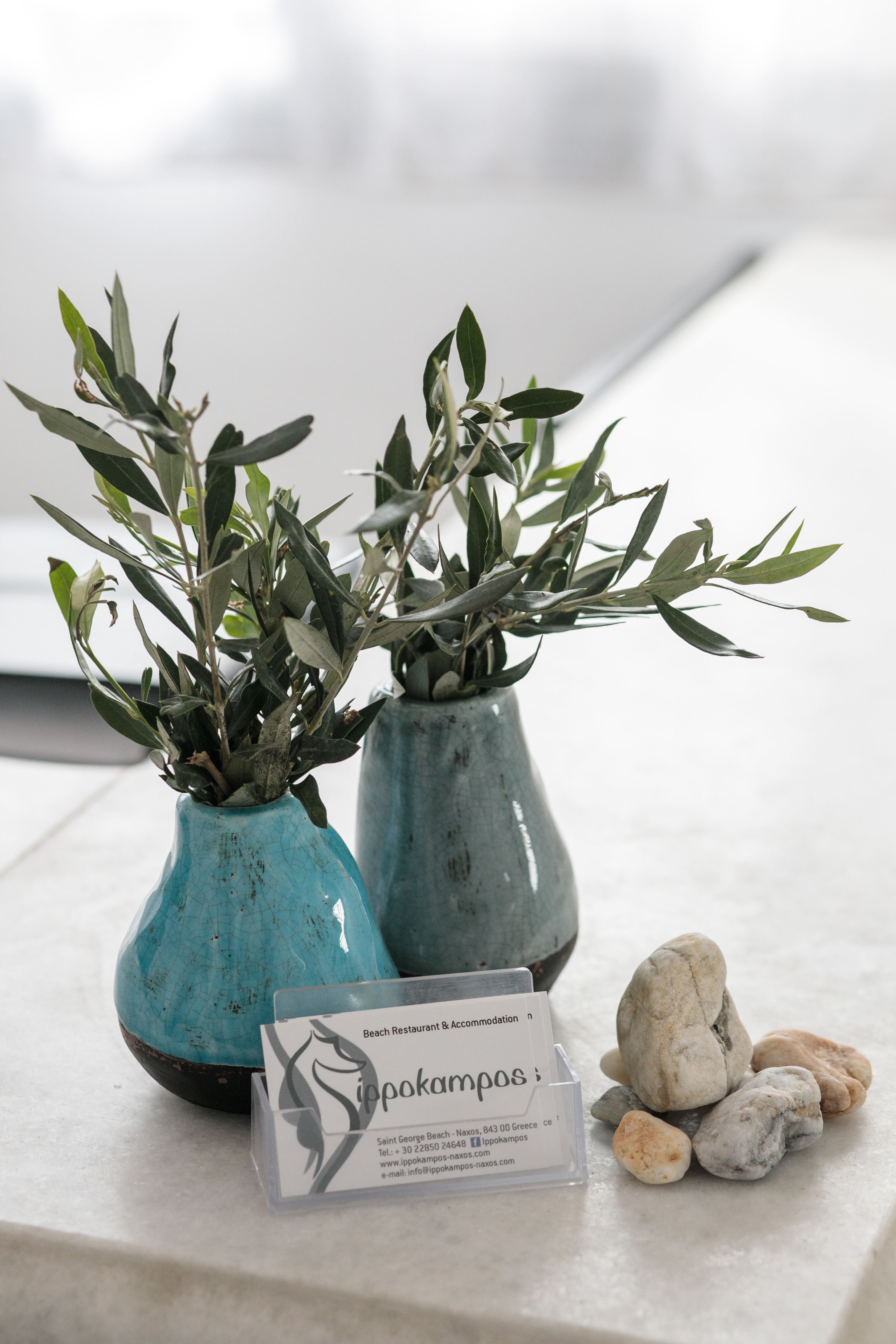 Two ceramic vases with olive branches on a table, a business card, and a pile of stones.