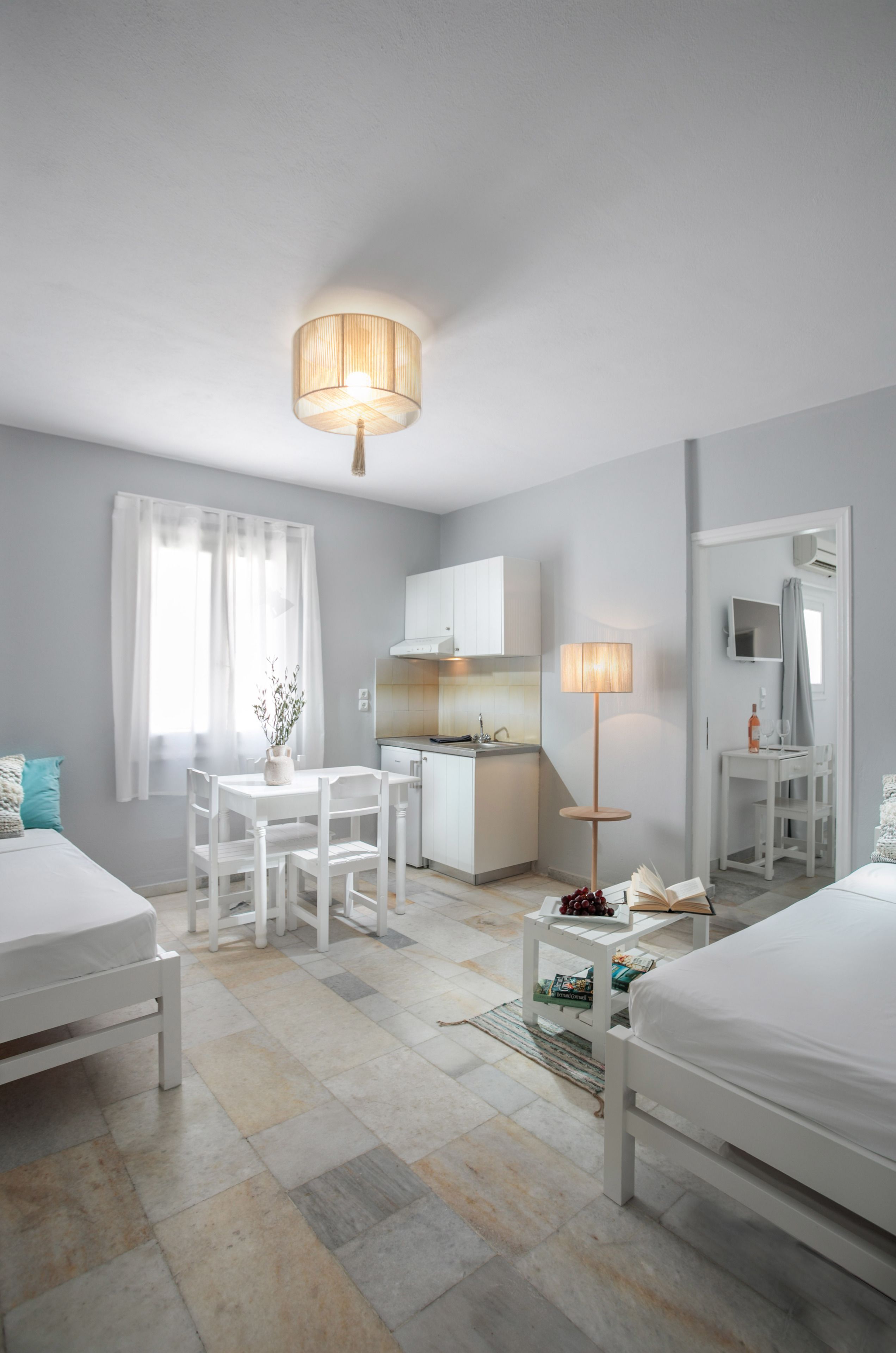 A modern hotel room with a bed, a desk, a chair, a small table with a lamp, and a kitchenette.
