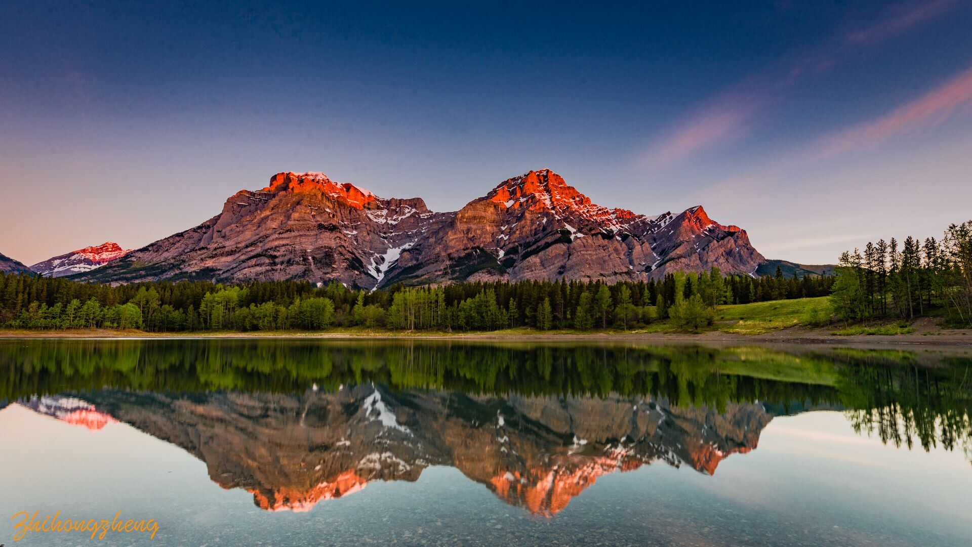 A mountain range with a clear reflection in a calm lake, surrounded by dense forest.