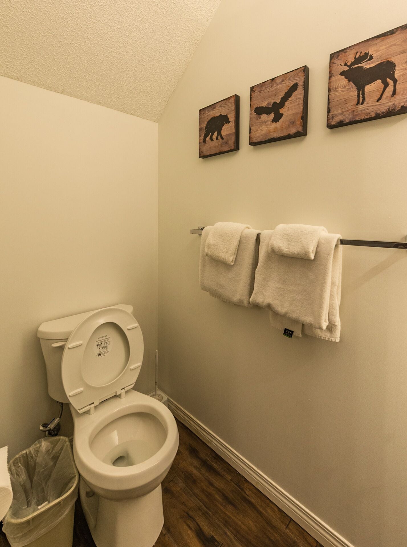 A bathroom with a toilet, a trash bin, and three wooden animal silhouette wall art pieces.