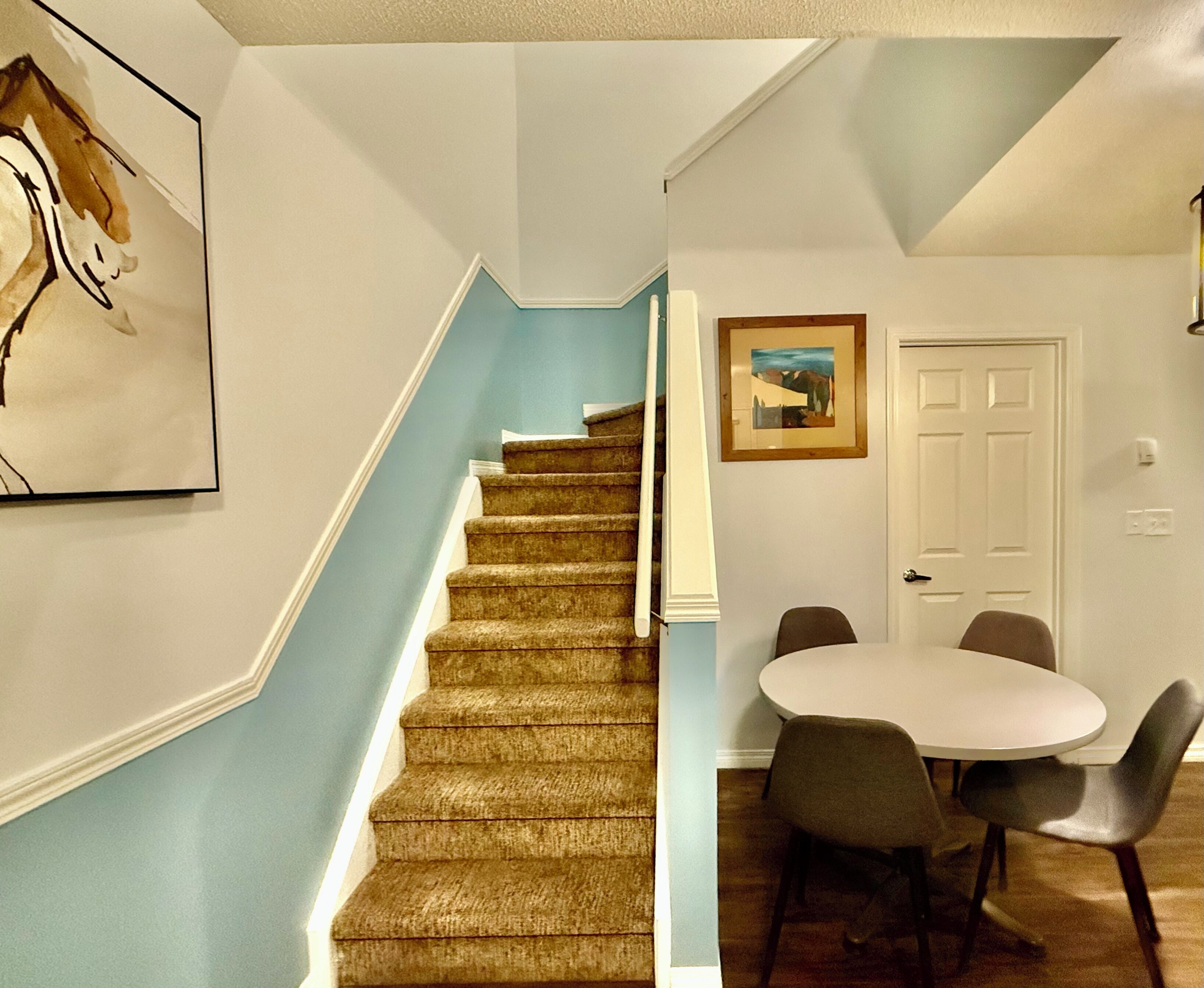 A staircase with a blue wall, a framed picture, and a dining area with chairs and a table.