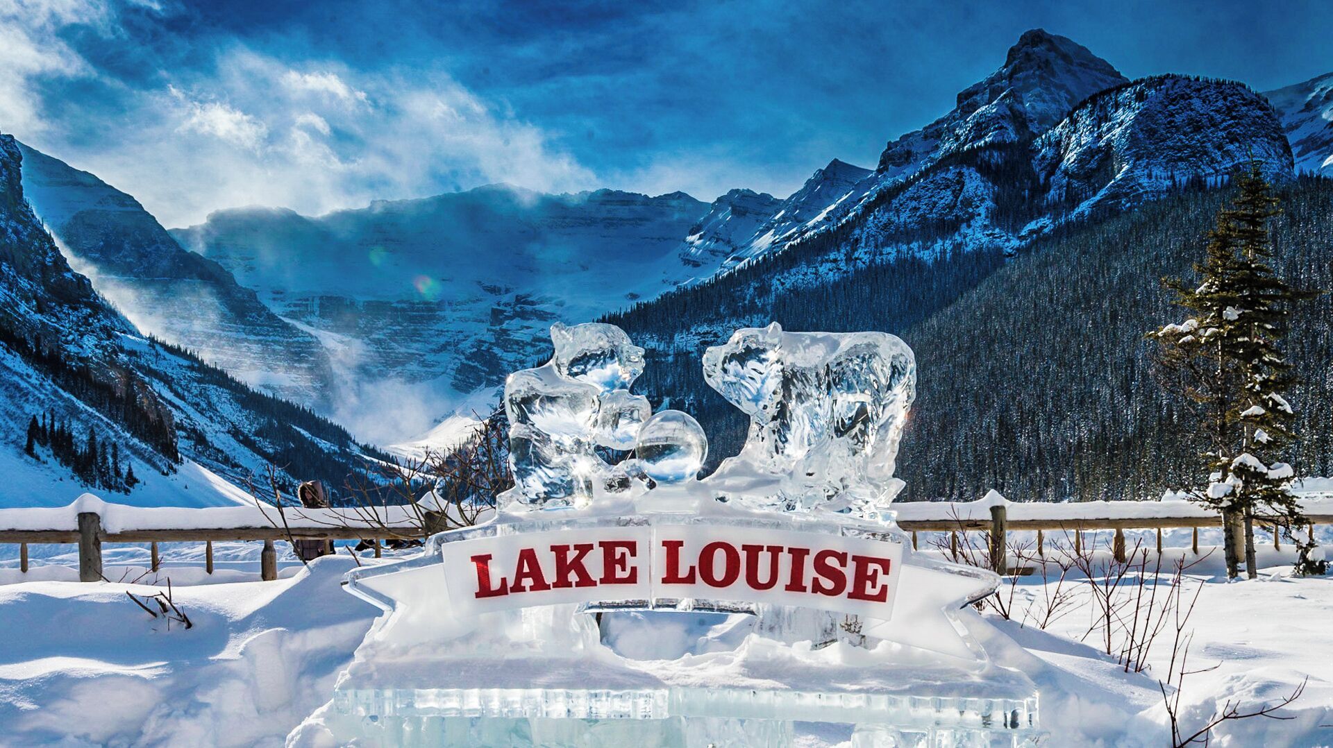An ice sculpture with 'LAKE LOUISE' text in the foreground, surrounded by snow-covered mountains and trees.