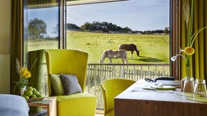 Una habitación con un ventanal grande, un sillón verde, un escritorio de madera y vistas a caballos pastando afuera.