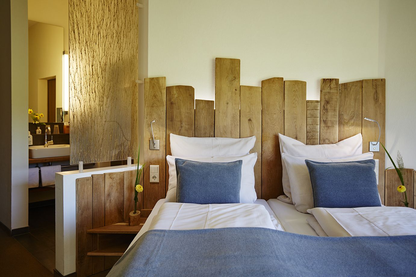 Une chambre à coucher avec une tête de lit en bois, deux lits avec du linge de lit blanc et des oreillers bleus, une table de chevet en bois et une salle de bain avec un lavabo et un miroir.